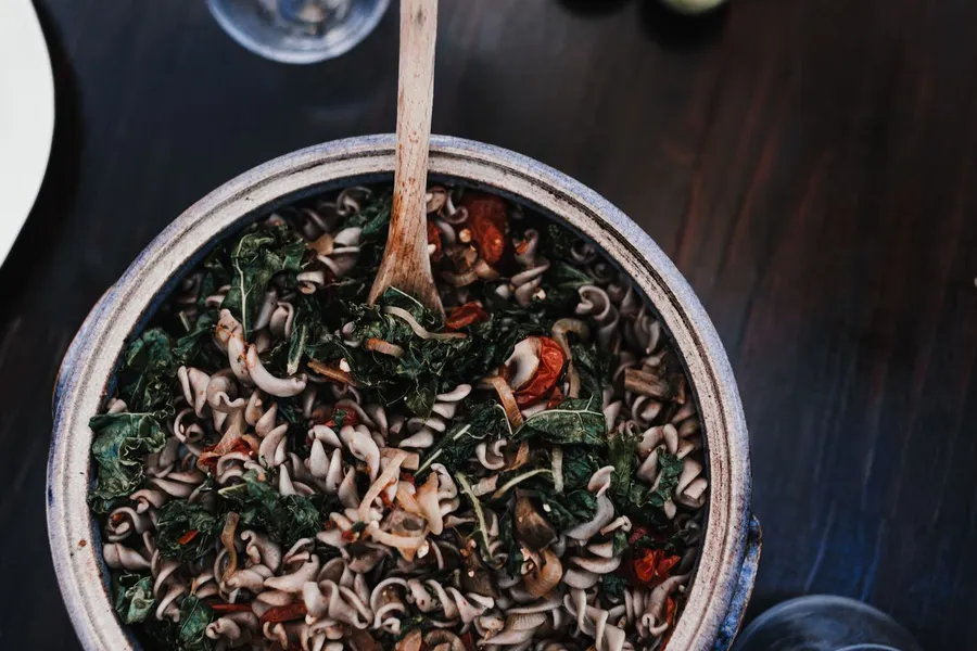 Top-down bowl of fusilli pasta salad with tomatoes, greens, and a wooden serving spoon