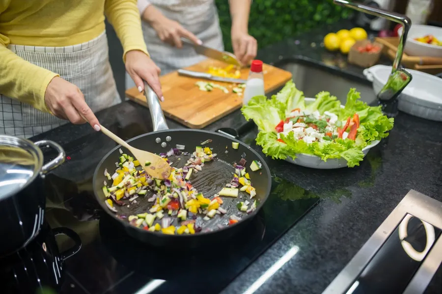 Home cooks stirring chopped vegetables in a wide skillet
